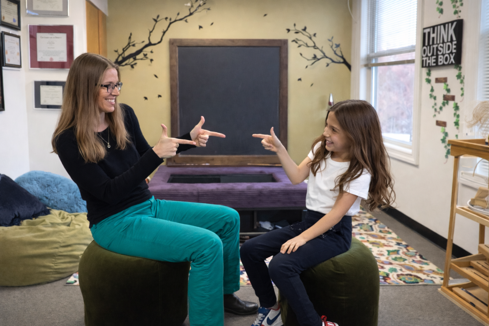 An adult and a child sit facing each other on round stools in a classroom, smiling and playfully pointing at one another during an interactive learning activity.