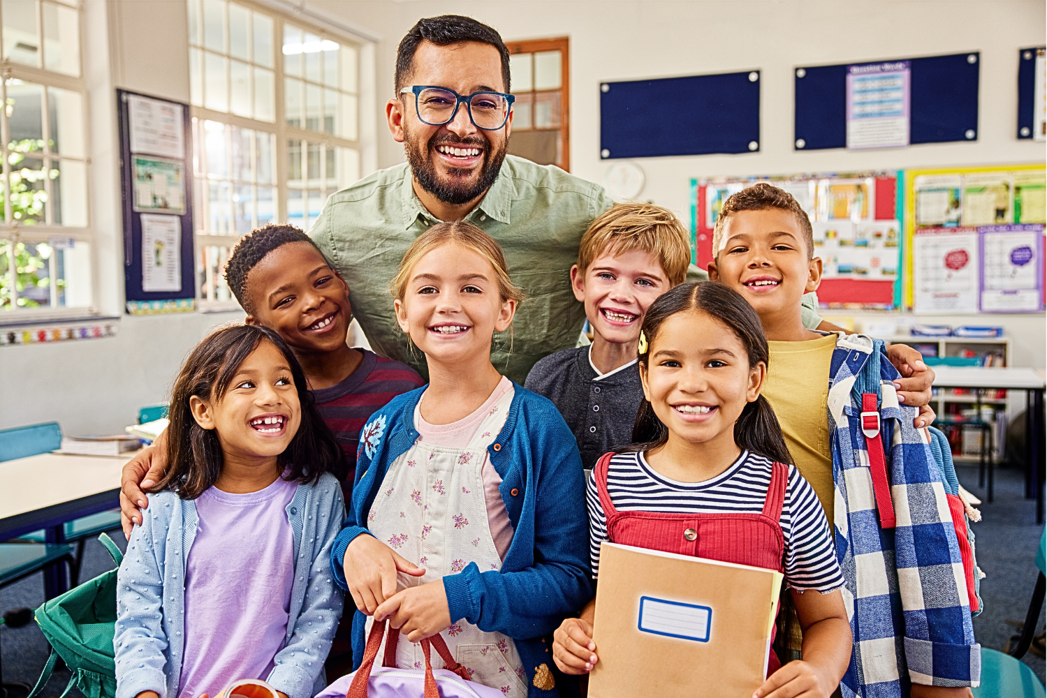A smiling teacher stands in a bright classroom surrounded by a diverse group of young students, who are smiling and holding school supplies.